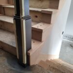 Black metal newel post installed at stair base with wood treads, risers, and skirt boards in raw finish.