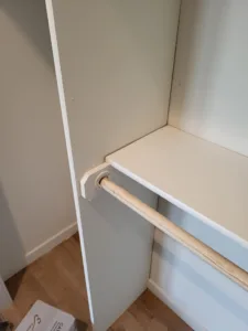 Closet shelf corner with painted white surround and unfinished natural wood hanging rod, wood floor below.