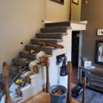 Staircase under construction with carpet treads, finished risers, newel posts, and handrail installed; work in progress with tools and materials visible.