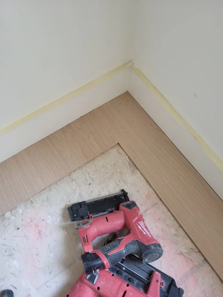 White baseboard pieces meeting at an inside wall corner, with a nailer resting on the protected floor.