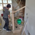 Carpenter installing curved handrail fitting on spiral staircase with wood balusters and newel post in progress.