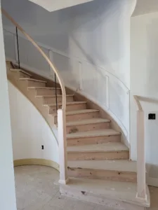 Curved wood handrail mounted to the wall above a staircase with raw wood treads and cable balusters.