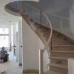 Spiral staircase with curved skirt board, wood treads, and glass railing—raw wood trim stage.