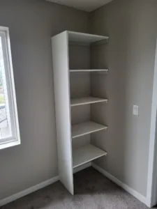 White painted corner closet shelving tower with five shelves, installed beside a window, carpet floor.