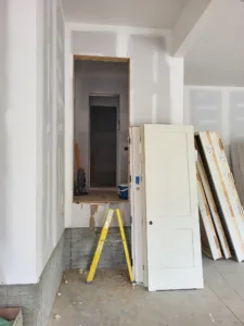 Hallway during door trim-out with a stepladder in the framed opening and panel door slabs staged nearby.