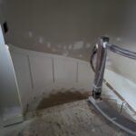 Newel post installation at curved staircase base with handrail in progress, drywall dust present.