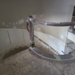 Curved staircase handrail and newel post installation in progress with protective wrapping and wainscoting panels.