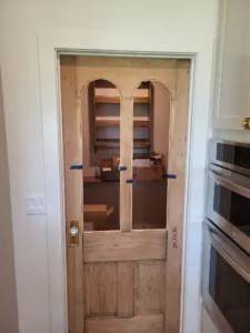 Unfinished wood door with arched glass panes and a paneled base, set in a cased opening