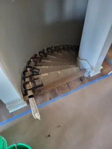 Laminated curved wood stair piece held in shape by many clamps, unfinished, resting on taped flooring