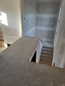 Interior stairwell viewed from above, open riser wood stairs below, subfloor and bare drywall walls.
