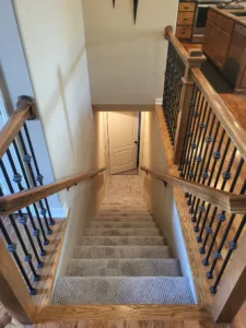 View down staircase with wood newels, iron balusters, carpet runner, and door at landing