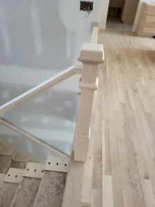 Wood newel post attached to a glass panel stair railing above light wood flooring