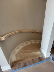 Curved wood handrail being installed along a staircase wall with finished treads and painted risers.