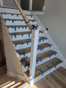 Staircase with finished wood treads, white risers, newel post, and handrail taped for paint protection.