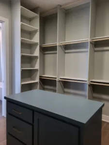 Walk-in closet with tiered corner shelving and cubbies, painted white, dark blue island dresser nearby.