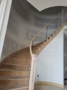 Wood newel post at base of a curved staircase, curved handrail and cable railing, treads unfinished.
