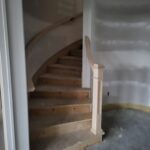 Newel post with curved handrail installed at base of spiral staircase; raw wood treads and risers visible.