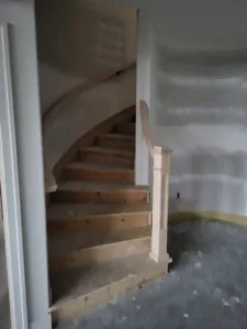 Newel post with a curved handrail at the base of an interior staircase, raw wood treads