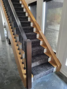 Carpeted staircase with wood posts, dark fabric runner, and vertical black metal balusters