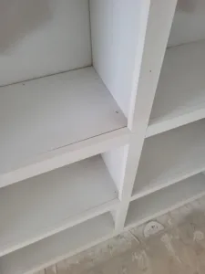 Painted white closet shelving corner with diagonal support brace and open shelf tiers, bare concrete floor.