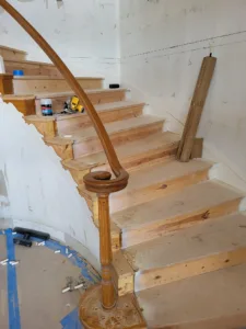 Curved staircase with a scalloped open stringer, turned newel post volute, and unfinished wood treads.