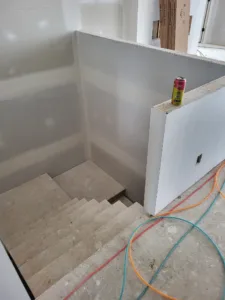Basement staircase under construction, drywalled walls, raw wood treads, wiring and hoses exposed nearby.