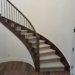 Curved staircase with dark wood skirt boards, newel posts, horizontal metal rod railing, and carpet treads installed.