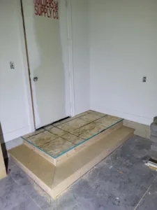Plywood platform under construction in corner nook, blue tape marking cut lines, concrete floor visible.