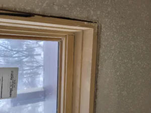 Raw wood window casing with mitered corner, unpainted, installed against a heavily textured interior wall.