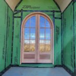 Arched-top exterior door slab with multi-pane glass installed during trim stage, raw wood finish, house wrap visible around opening.