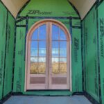 Arched-top exterior door with dual glass panes and wood jamb installed in rough opening with house wrap and tape.
