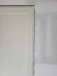 Painted white door slab set in jamb with casing partly installed, raw trim visible at top corner
