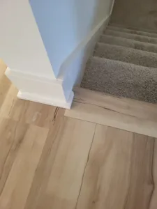 Painted white baseboard at a stair newel base, hardwood flooring meeting carpeted stairs beyond