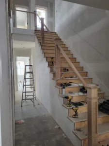 Unfinished wood staircase along a hallway wall with newel post installed and tools staged on treads.