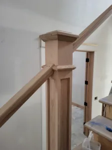 Unfinished wood newel post with flat cap connecting to stair handrail, drywall not yet painted