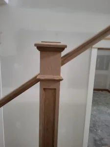 Unfinished wood newel post with cap and base trim supporting a stair handrail, set against drywall