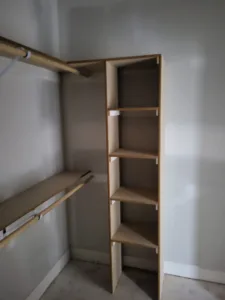 Closet corner with natural wood hanging rod and small vertical shelf ladder unit, unfinished raw wood.