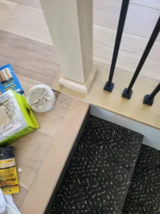 Iron stair balusters mounted in black bases on a wood cap, with wood filler staged nearby.