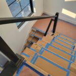 Black metal newel post and handrail installed on staircase with raw wood treads taped for protection during construction.