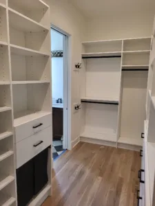 Completed walk-in closet with white shelving, drawers, and hanging rods, black hardware, wood floor.