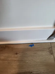 Painted white baseboard installed above wood-look flooring with painter's tape on the floor