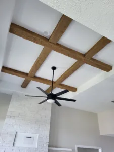 Dark stained wood ceiling beams crossing in an X pattern above a fan and stone fireplace