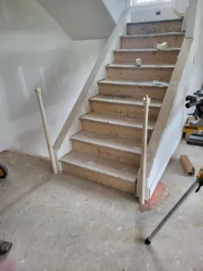 Unfinished stair skirt boards and newel post installed on interior staircase, drywalled walls behind.