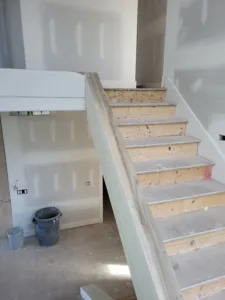 Interior stair in progress with bare tread blanks beside a drywall half wall topped with a wide wood cap