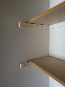 Two particleboard closet shelves with bullnose edges mounted on wood cleats, unfinished raw wood.