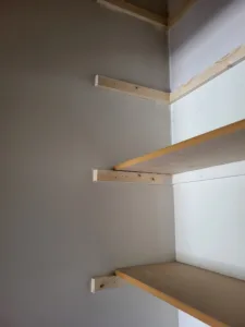 Three particle board closet shelves on wood cleats being installed at a corner, raw and unfinished.