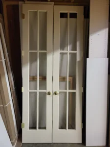 White primed French doors with multi-lite glass panels and brass hinges, staged before installation.