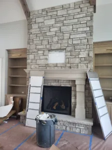 Stone veneer fireplace with carved stone mantel shelf, under construction, flanked by built-in wood shelving.