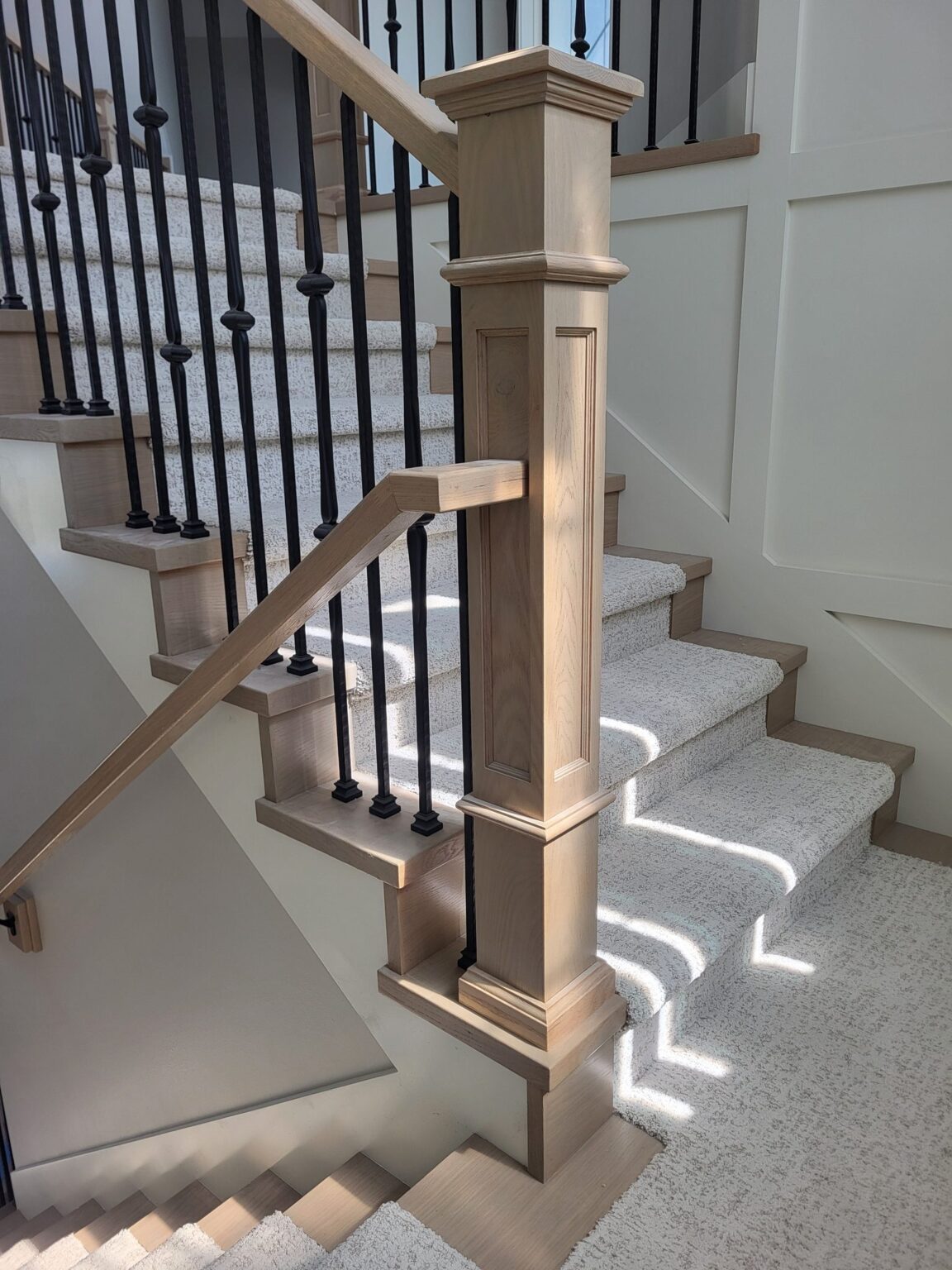 Stained wood newel post and wrought-iron balusters against a white board-and-batten stairwell wall, sunlight across the carpeted stairs