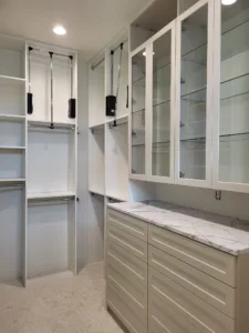 Finished white closet cabinetry with glass-front uppers, pull-down hanging rods, and marble countertop.